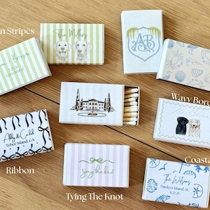 May include: A collection of decorative matchboxes with various designs. Some boxes feature text like "Thin Stripes," "The Millers," and "Abby & Caleb." Other designs include illustrations of dogs, coastal themes, and a ribbon motif. The matchboxes are on a wooden surface.