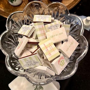 May include: A clear, scalloped glass bowl filled with small matchboxes. The matchboxes have various designs, including striped patterns, floral motifs, and text that reads "April 18, 2026" and "The Thompson's".