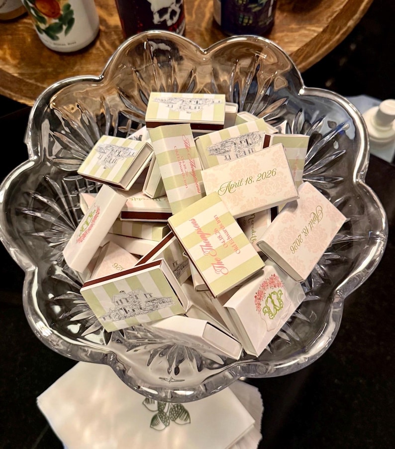 May include: A clear, scalloped glass bowl filled with small matchboxes. The matchboxes have various designs, including striped patterns, floral motifs, and text that reads "April 18, 2026" and "The Thompson's".