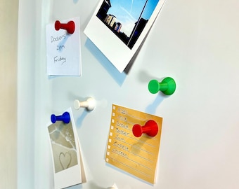 May include: A collection of colorful magnetic push pins in green, blue, red, and white, holding various notes and photos on a white surface. The notes include a doctor's appointment and a grocery list. The photos feature a beach scene, a bouquet, and a landscape.