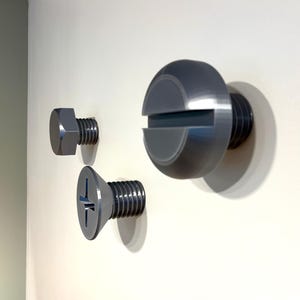 Puede incluir: Tres tornillos metálicos grises de diferentes tamaños están montados en una pared blanca. Los tornillos incluyen una cabeza hexagonal, una cabeza plana y una cabeza ranurada grande. Los tornillos están dispuestos en línea horizontal.