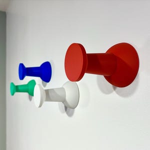 May include: Four brightly coloured wall hooks in red, blue, green, and white. Each hook features a circular head and a cylindrical body, mounted on a white wall. These hooks are designed for hanging items.