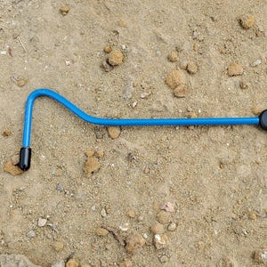 May include: A blue and black tool with a curved end and a handle. The tool is lying on a sandy surface. The handle is black and has a hole for hanging.