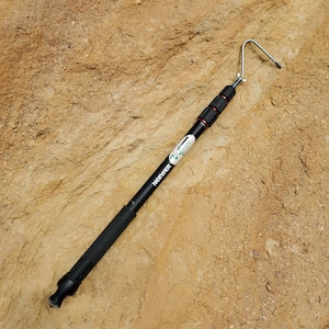 May include: A black NEEWER telescoping pole with a silver hook at the top. The pole has red accents and a textured grip. The pole is lying on a textured, light brown surface.