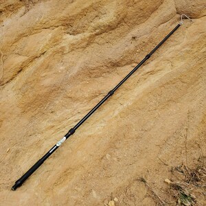 May include: A black telescopic pole with a hook, possibly for photography or outdoor activities. The pole is extended and lies on a textured, light brown rock surface. The brand name "Neewer" is visible on the pole.