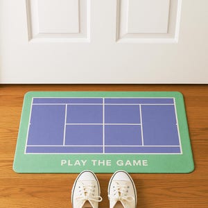 Puede incluir: Una alfombra rectangular con un diseño de cancha de tenis. El área de juego es azul con líneas blancas, y el borde es verde. Las palabras "PLAY THE GAME" están impresas en blanco. Zapatillas blancas en la parte inferior.