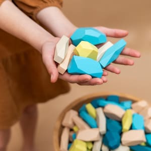 May include: A person's hands holding a selection of colourful wooden geometric blocks. The blocks are in shades of blue, yellow, and beige. More blocks are in a wooden bowl below. A fun toy.