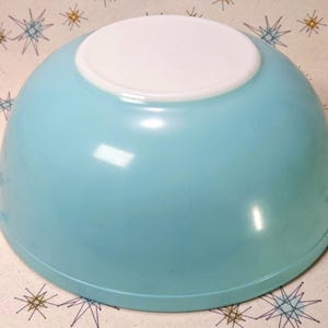 May include: A round mixing bowl with a smooth, glossy finish. The exterior of the bowl is a solid, vibrant light blue color, while the base and the underside are a contrasting, solid white. The bowl has a wide, open top and a curved shape.