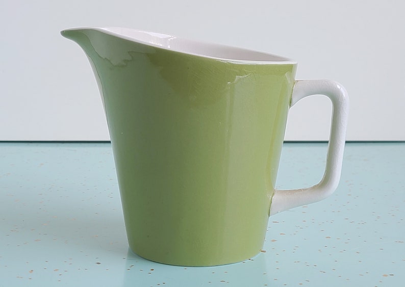 Puede incluir: Una jarra de cer&aacute;mica verde con asa blanca y interior blanco. La jarra tiene una forma redondeada ligeramente acampanada.