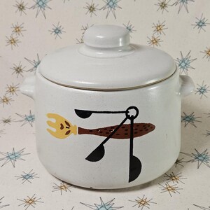 May include: A white ceramic jar with a lid and two small handles. The jar features a whimsical design of a yellow whisk, brown spatula, and black measuring spoons. The jar is likely for kitchen use.