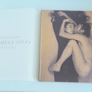 May include: A black and white photo book titled "Annie Leibovitz 1970-1980 Photographs". The cover features a black and white photograph of two people embracing. The woman is on top of the man, her arms wrapped around his neck. The man's arm is around her waist. The woman's hair is long and flowing. The man's hair is short. The photo is in a soft focus.