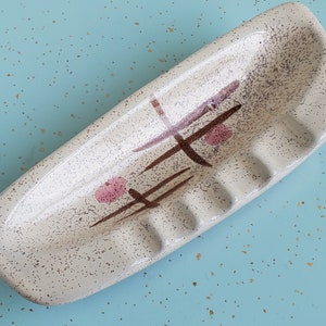 May include: A white ceramic ashtray with a brown and pink abstract design. The ashtray has five indentations for cigarettes.