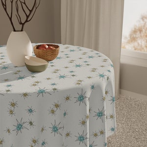 May include: A white round tablecloth with a repeating pattern of teal and gold starbursts. The tablecloth is on a round table with a bowl of fruit and a vase with branches.