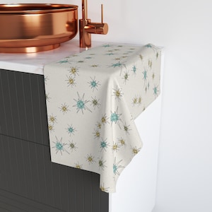 May include: A white table runner with a pattern of teal and yellow starbursts. The runner is draped over a countertop with a copper sink.