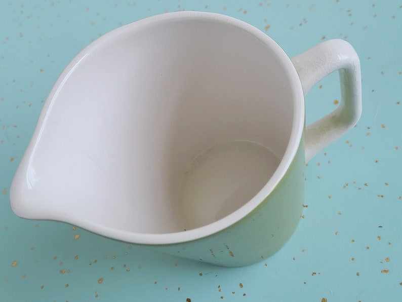 Puede incluir: Una jarra de cer&aacute;mica verde con interior blanco y asa. La jarra est&aacute; vac&iacute;a y tiene un ligero labio en el lado del vertido.