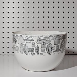 May include: White ceramic bowl with a black and white mushroom illustration around the rim. The bowl has a rounded shape and sits on a white surface. The background is a white pegboard with black dots.