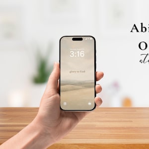 Puede incluir: Un teléfono inteligente negro muestra un fondo de pantalla de paisaje con el texto "3:16" y "glory to God". El teléfono se sostiene contra un fondo borroso con el texto "Abiding Olive studio."