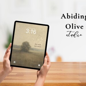 Puede incluir: Una tableta negra que muestra una imagen de paisaje con el texto "3:16" y "His will, not mine". La tableta está sostenida por manos sobre una superficie de madera. Las palabras "Abiding Olive studio" están escritas en escritura negra.