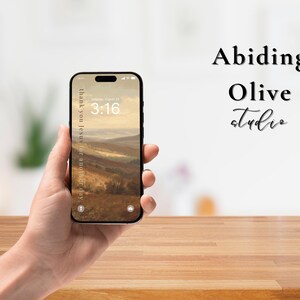 May include: A black smartphone displays a landscape image with the text "Thank you Jesus for another day" and "3:16". The phone is held in a hand, with the words "Abiding Olive Studio" in the background.