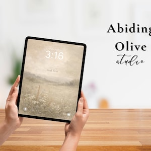 May include: A black tablet displaying a serene landscape wallpaper with the time 3:16 and the words "God first." The tablet is held by hands over a wooden surface. The words "Abiding Olive Studio" are written in black.