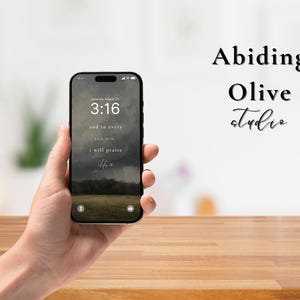 May include: A black smartphone displaying a landscape wallpaper with the text "3:16" and inspirational text. The phone is held in a hand, with the words "Abiding Olive studio" in the background.