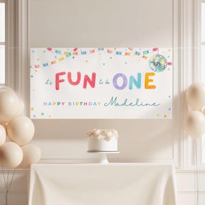 May include: A white banner with the words "FUN ONE" in colorful letters, with the text "Happy Birthday Madeline" below. The banner is decorated with a string of colorful lights, stars, and a disco ball. A white cake sits on a table.