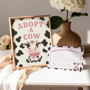 Può includere: Una stampa incorniciata con la scritta "ADOTTA UNA MUCCA" e un'illustrazione di una mucca dei cartoni animati. Un certificato di adozione abbinato è accanto alla cornice. Lo sfondo presenta macchie nere di mucca.
