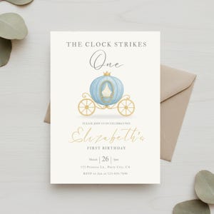 Op de afbeelding: Crèmekleurige uitnodigingskaart met een illustratie van een blauwe koets en gouden accenten. De tekst luidt "The Clock Strikes One" en "Elizabeth's First Birthday" met datum en tijd. Uitnodiging voor een eerste verjaardag.
