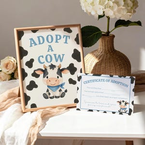 May include: A framed print with the text "ADOPT A COW" and a cartoon cow illustration. A matching certificate of adoption is next to the frame. A vase with white flowers and a beige table complete the scene.