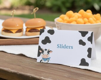 May include: A wooden table holds a white card with the word "Sliders" and a cartoon cow illustration. Two mini cheeseburgers with toothpicks sit on a wooden tray, and a bowl of orange snacks is in the background.