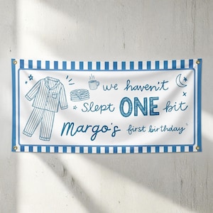 以下が含まれることがあります： 青いストライプと「we haven't slept ONE bit Margo's first birthday」というテキストが入った白いバナー。バナーには、パジャマ、パンケーキ、コーヒーカップ、月、星のイラストが描かれています。