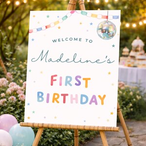 May include: A white sign on a wooden easel, reading "Welcome to Madeline's First Birthday" in colourful letters. The sign is decorated with stars, a disco ball, and a garland of shapes. Balloons are in the foreground, with a garden backdrop.