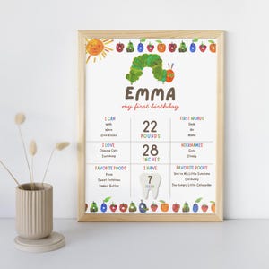 May include: A framed print for a first birthday, featuring the name "EMMA" and details like weight (22 pounds), height (28 inches), and favorite things. The design includes a sun, a caterpillar, and colorful fruits.