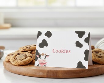 May include: A wooden serving board holds several chocolate chip cookies. A white card with black cow spots and the word "Cookies" in pink features a cartoon cow with a pink scarf. The cookies are arranged around the card.