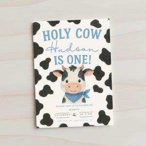 May include: A rectangular invitation card with a cow theme. The text "HOLY COW Hudson IS ONE!" is written in blue and black. A cartoon cow illustration is in the center. The card also includes event details: date, time, and location.