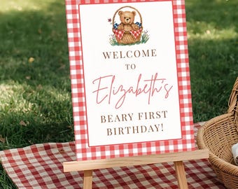 May include: A red and white gingham-patterned sign on a wooden easel. The sign reads "Welcome to Elizabeth's Beary First Birthday!" and features a teddy bear in a basket. The sign is set on a red and white gingham picnic blanket.