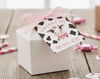 May include: A white gift box tied with a pink ribbon and a tag that says "Thanks so Moo-ch". The tag features a cartoon cow with black spots and a pink bandana. The box is on a wooden surface.