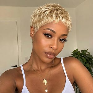 Peut inclure: Une perruque courte de style pixie blond texturé avec des couches ondulées. Les cheveux sont d'une teinte blond platine vif avec des racines plus foncées. La coupe présente une longueur courte qui encadre le visage, avec une ligne de cheveux naturelle.
