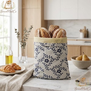 May include: A fabric bread bag with a blue floral pattern, holding several loaves of bread. The bag has a yellow interior and sits on a white countertop. Croissants and pears are also visible.