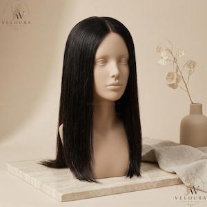 May include: A black wig with long, straight hair is displayed on a mannequin head. The wig is centered on a light-colored, rectangular surface. The background is a neutral beige, with a small vase and decorative plant to the right.