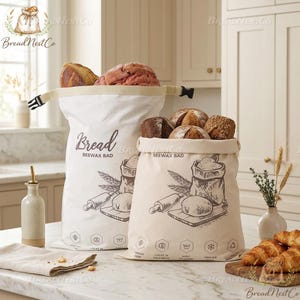 Puede incluir: Dos bolsas de pan de lona llenas de hogazas de pan. Las bolsas tienen un cierre de cordón y presentan un gráfico de herramientas para hacer pan. El texto "Bread BEEWAX BAD" está impreso en las bolsas. También se ve una botella de aceite de oliva y una tabla de cortar de madera con cruasanes.