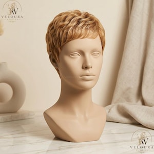 Puede incluir: Una cabeza de maniquí con una peluca corta y ondulada de color marrón claro. El maniquí es de color beige neutro, con una superficie lisa y rasgos faciales definidos. La peluca tiene un aspecto y una textura naturales. El logotipo de Veloura Wigs Co. está en la esquina superior izquierda e inferior derecha.
