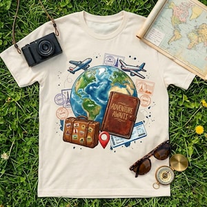 May include: Cream-colored t-shirt featuring a travel-themed graphic. The design includes a globe, airplanes, a suitcase, a journal with "Adventure Awaits" text, and travel stamps. A camera, sunglasses, and a compass are also visible.