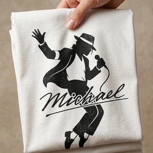 Puede incluir: Una camiseta blanca doblada con una silueta negra de un artista con sombrero y traje, sosteniendo un micr&oacute;fono. La palabra "Michael" est&aacute; escrita en cursiva debajo.