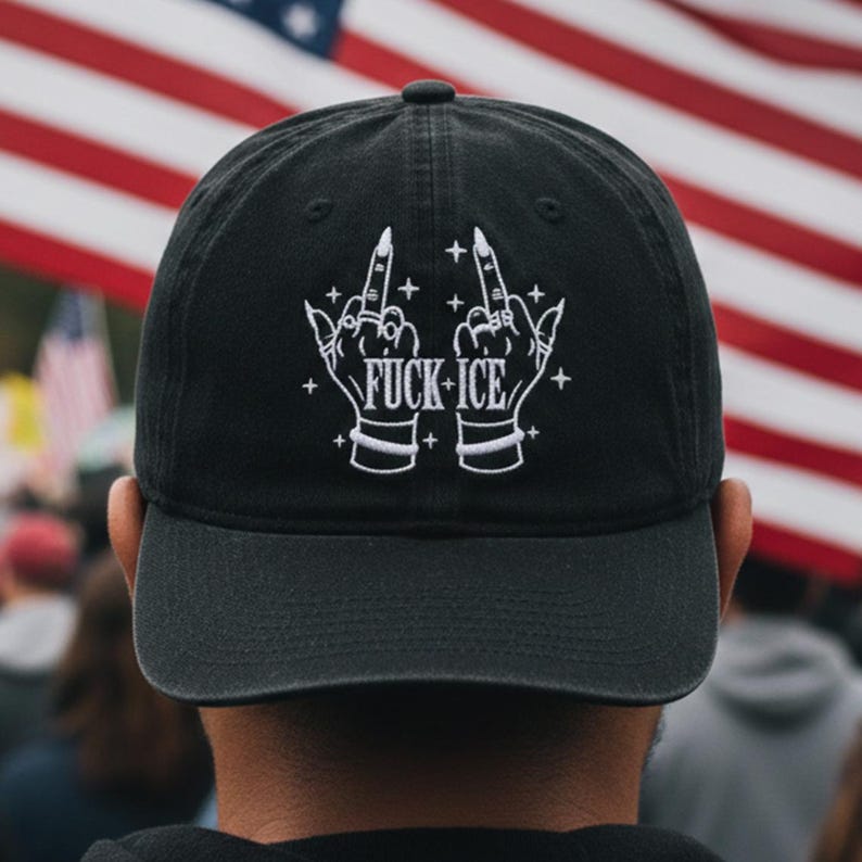 May include: Black baseball cap featuring the embroidered text "FUCK ICE" in white, accompanied by two hands giving the middle finger. The cap is worn by a person, with the American flag visible in the background.