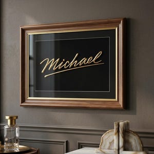 Puede incluir: Una obra de arte enmarcada con el nombre "Michael" en escritura dorada sobre un fondo negro. El marco es de madera oscura con un borde interior dorado. La obra de arte se exhibe en una pared gris.