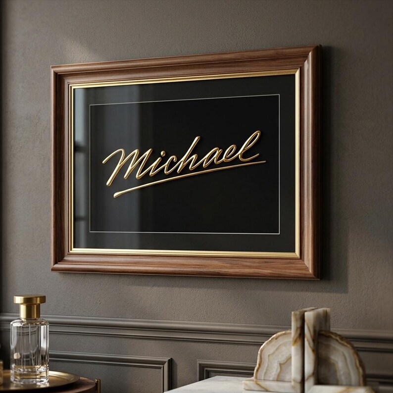 Puede incluir: Una obra de arte enmarcada con el nombre "Michael" en escritura dorada sobre un fondo negro. El marco es de madera oscura con un borde interior dorado. La obra de arte se exhibe en una pared gris.
