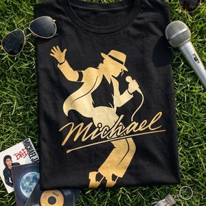 Puede incluir: Camiseta negra con una silueta dorada de un artista con micr&oacute;fono y sombrero, y el nombre "Michael" en cursiva. Gafas de sol, un micr&oacute;fono y discos de vinilo est&aacute;n colocados sobre hierba alrededor de la camiseta.