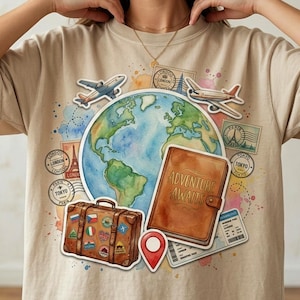 May include: Beige t-shirt with a travel-themed graphic. The design includes a watercolor globe, airplanes, a suitcase, stamps, a journal with "ADVENTURE AWAITS" text, and a red location pin. The shirt is a casual wear item.