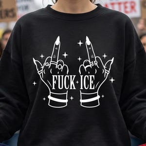 May include: Black sweatshirt featuring a white graphic of two hands displaying the middle finger, with the text "FUCK ICE". The hands are decorated with rings and bracelets, and surrounded by star shapes.